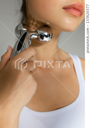 Close-up of a young woman massaging her neck with a metal 3D roller, demonstrating a tightening and relaxing skincare routine at home. Close-up of a young woman massaging her neck with a metal 3D roller, demonstrating a tightening and relaxing skincare routine at home. 133620377