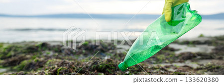 Volunteer picking up a green plastic bottle during beach cleanup, highlighting pollution, waste removal, and environmental protection. 133620380