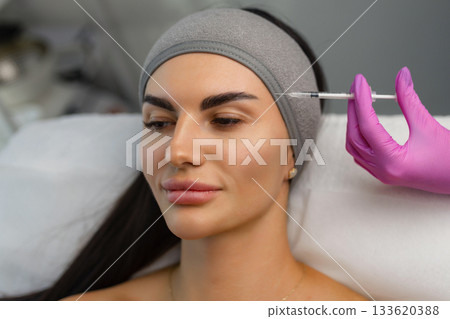 Close up of hands of young cosmetologist injecting botox in female face. She is standing and smiling. The woman is closed her eyes with relaxation 133620388
