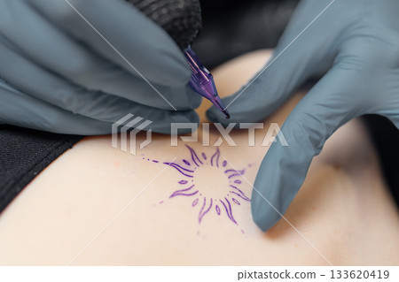 close-up of a tattoo artist outlining a sun design on a client skin, showing the precision of the tattoo process, sterile gloves, and professional equipment. close-up of a tattoo artist outlining a sun design on a client skin, showing the precision of the tattoo process, sterile gloves, and professional equipment. 133620419