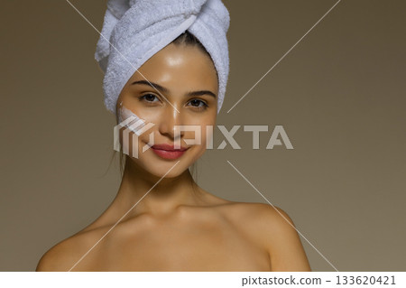 Young woman applying skincare cream to her cheek after a shower, wearing a towel on her head and showing a gentle beauty routine. Young woman applying skincare cream to her cheek after a shower, wearing a towel on her head and showing a gentle beauty routine. 133620421