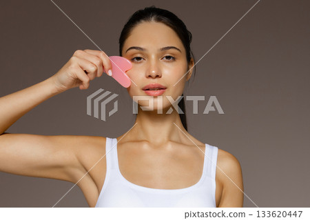 Young woman gently massaging her face with a pink gua sha stone, demonstrating a skincare routine against a neutral background. Young woman gently massaging her face with a pink gua sha stone, demonstrating a skincare routine against a neutral background. 133620447