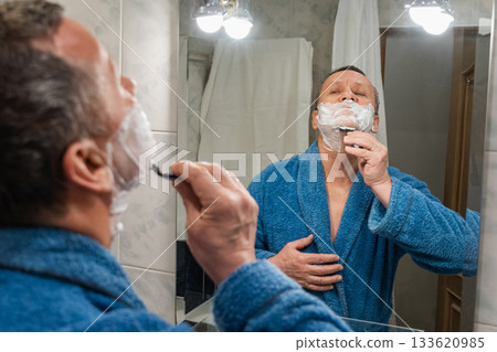 Man Shaving wearing blue blue robe in Bathroom Mirror. Morning Shaving Routine Man Shaving wearing blue blue robe in Bathroom Mirror. Morning Shaving Routine 133620985