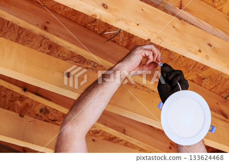Electrician is fixing lighting fixture to ceiling in home renovation setting, on details safety. Electrician is fixing lighting fixture to ceiling in home renovation setting, on details safety. 133624468
