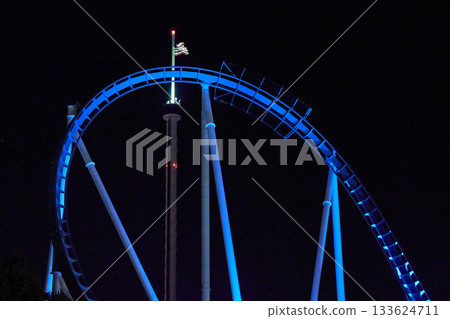 A blue steel roller coaster curves high against a dark sky while an illuminated tower and flag rise behind it. Bright lights outline the track and supports, creating a dramatic night scene. A blue steel roller coaster curves high against a dark sky while an illuminated tower and flag rise behind it. Bright lights outline the track and supports, creating a dramatic night scene. 133624711