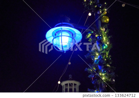 A blue lantern glows beside Christmas garland and ornaments at night. A soft lighthouse silhouette and string lights complete the coastal holiday scene. 133624712