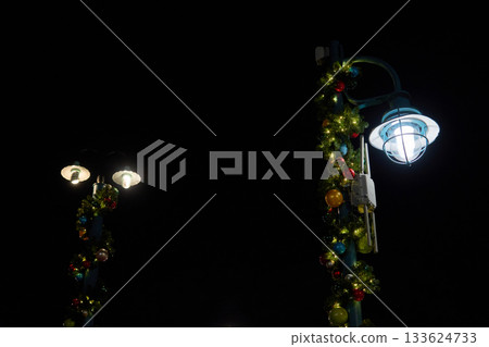 A pair of street lamps decorated with Christmas garland and colorful ornaments glow against a dark sky. The scene shows simple holiday lighting on a quiet walkway in a theme park. 133624733