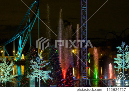 Tall water jets and fountains glow with red, green, blue, and purple lights during a Christmas show in a theme park at night. Decorated trees and a roller coaster frame the vibrant holiday scene. 133624737