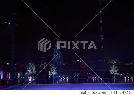 A towering blue light tree stands across the lagoon with fountain arcs and reflections. The ice stage and holiday decorations glow under the dark sky. A towering blue light tree stands across the lagoon with fountain arcs and reflections. The ice stage and holiday decorations glow under the dark sky. 133624746