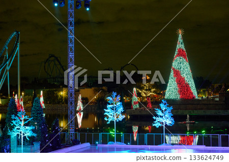 A towering red and white light tree glows across the lagoon next to a bright ice rink. Spotlights, coaster tracks, and floating trees frame the festive scene. A towering red and white light tree glows across the lagoon next to a bright ice rink. Spotlights, coaster tracks, and floating trees frame the festive scene. 133624749