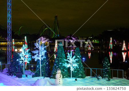 Artificial snow, glowing trees, and a snowman decorate a theme park stage beside a lagoon. Roller coaster loops and floating light trees shine in the dark sky. Artificial snow, glowing trees, and a snowman decorate a theme park stage beside a lagoon. Roller coaster loops and floating light trees shine in the dark sky. 133624750