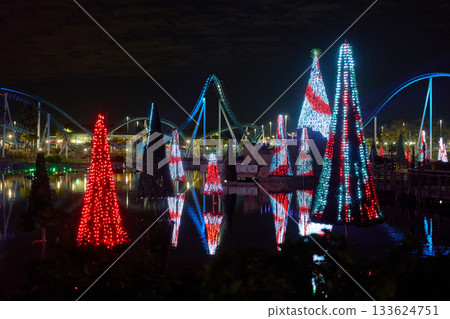 Striped red and white Christmas light trees float on a lagoon with vivid reflections. Curved coaster tracks and park lights create a bright winter night panorama. Striped red and white Christmas light trees float on a lagoon with vivid reflections. Curved coaster tracks and park lights create a bright winter night panorama. 133624751