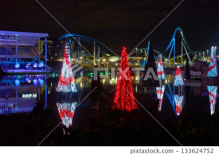Conical red and white light trees shimmer on the water with perfect mirror reflections. A looping roller coaster frames the bright holiday scene at night. 133624752