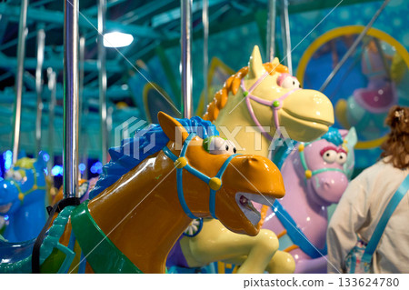 Close view of a bright cartoon carousel horse with a blue mane and shiny bridle. Vibrant lights and other ride figures glow in the background. 133624780
