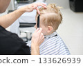 Barbershop stylist combing blonde child hair for haircut 133626939