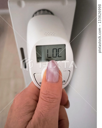 A woman's hand presses a button to adjust a modern thermostat on a white radiator. A woman's hand presses a button to adjust a modern thermostat on a white radiator. 133626998