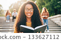 Young woman student with curly hair is reading a book outdoors on steps, surrounded by peers, showcasing a moment of learning and engagement in education 133627112