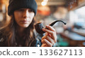 Woman in black beanie is thoughtfully holding a smoking pipe, surrounded by a cozy cafe atmosphere, reflecting on moments of contemplation 133627113