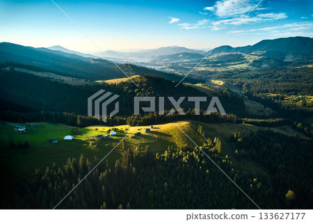 Aerial view of picturesque valley with rolling hills, lush forests, and scattered houses. Sunlight bathes landscape in warm glow, highlighting vibrant greens and creating serene atmosphere. 133627177