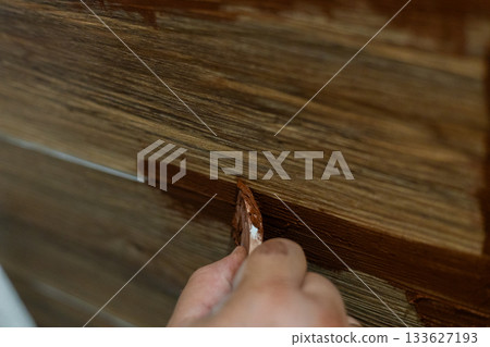 A close-up view of a person's hand using a putty knife to apply brown grout between the joints of wood-look ceramic floor tiles. The man is doing DIY home improvement and renovation work. A close-up view of a person's hand using a putty knife to apply brown grout between the joints of wood-look ceramic floor tiles. The man is doing DIY home improvement and renovation work. 133627193