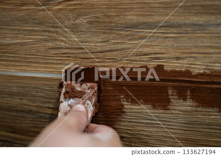 Applying brown grout by hand with a trowel to fill narrow joints between wood-look ceramic tiles during a renovation. Close-up of the wall renovation and finishing work. 133627194