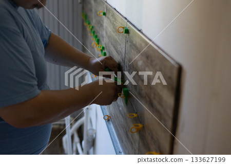 Man using tile leveling tools to install wood-look ceramic wall tiles in a bathroom renovation, close-up of skilled hands ensuring precise, even tile placement and finish. Man using tile leveling tools to install wood-look ceramic wall tiles in a bathroom renovation, close-up of skilled hands ensuring precise, even tile placement and finish. 133627199