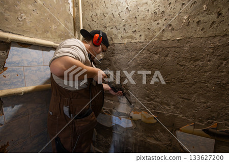 A worker in protective clothing operates a hammer drill to remove old plaster and wall tiles. A man uses a hammer drill for demolition work during a home or bathroom renovation project. 133627200