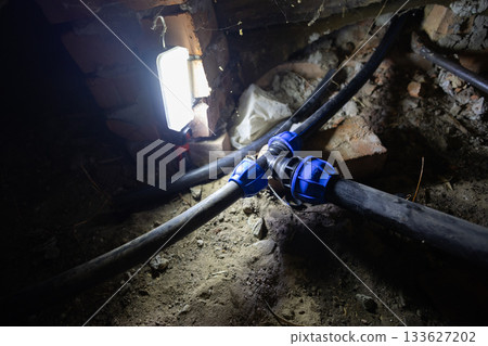 Basement water pipes with a black plastic adapter joining multiple lines, lit by a work light during repair or installation, showing close-up plumbing maintenance in a home. Basement water pipes with a black plastic adapter joining multiple lines, lit by a work light during repair or installation, showing close-up plumbing maintenance in a home. 133627202