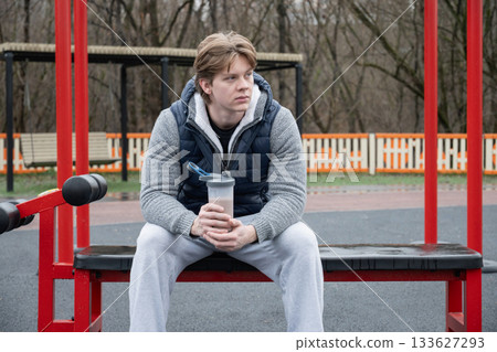Young man sits on an outdoor gym bench holding protein shake, wearing casual vest. He relaxed, enjoying moment of rest during workout. 133627293