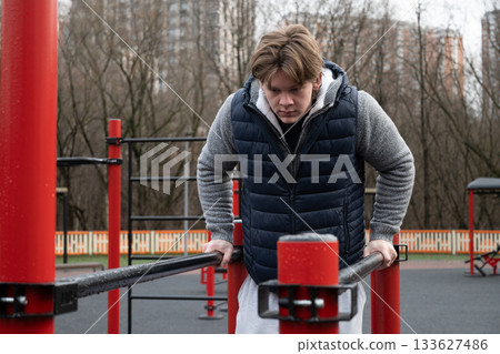 Focused man exercise on parallel bars in an urban outdoor gym after rain. Dressed warmly, concentrating on fitness routine amidst city backdrop, emphasizing strength and determination. Daily routine 133627486
