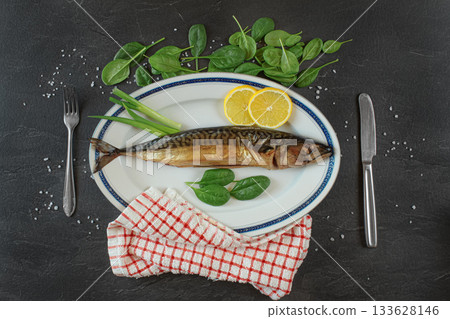 Whole smoked mackerel fish on white oval plate, gray black stone like table under, rock salt, lemon slices and green leaves salad near. View from above 133628146