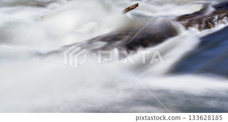Long exposure photo - water flowing over rock near drifted tree branch, everything smooth, only stationary wood and rock is in focus Long exposure photo - water flowing over rock near drifted tree branch, everything smooth, only stationary wood and rock is in focus 133628185