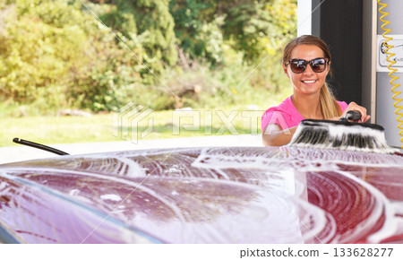 Young woman wearing pink t shirt and sunglasses cleaning her car in self serve carwash, blurred sun lit trees background 133628277