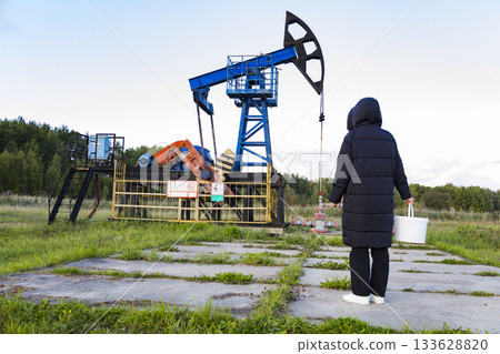 Worker performs maintenance, Technician checks machinery outdoors, Field staff inspects oil extraction device, Laborer surveys industrial equipment amidst cloudy environment 133628820