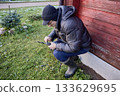 Homeowner replaces drill bit in power drill chuck while squatting near wall of house. 133629695