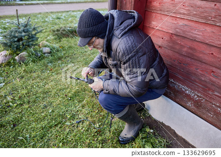 Homeowner replaces drill bit in power drill chuck while squatting near wall of house. 133629695