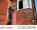 Drilling hole for surveillance camera cable, technician mounts device on facade of rural house, worker stands on ladder near window. 133629696