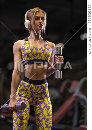 girl stands at the gym focused on her workout while doing bicep curls with shiny dumbbells. She wears colorful workout clothes and headphones highlighting her determination. 133630132