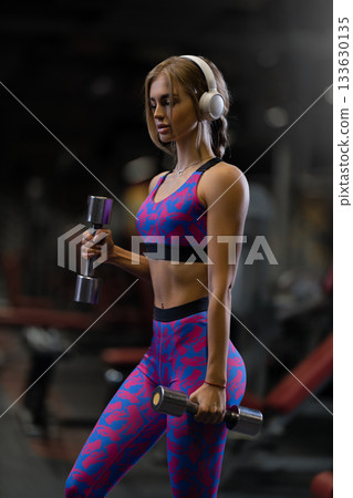 girl focuses on her form while doing bicep curls with shiny dumbbells. She wears workout clothes and headphones in a well lit gym showing determination and strength. girl focuses on her form while doing bicep curls with shiny dumbbells. She wears workout clothes and headphones in a well lit gym showing determination and strength. 133630135