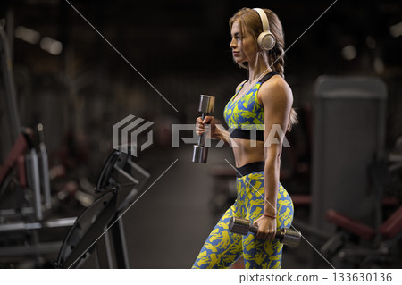 girl focuses on her workout routine in a gym doing bicep curls with shiny dumbbells. She wears a colorful outfit and headphones showcasing her determination and strength. girl focuses on her workout routine in a gym doing bicep curls with shiny dumbbells. She wears a colorful outfit and headphones showcasing her determination and strength. 133630136
