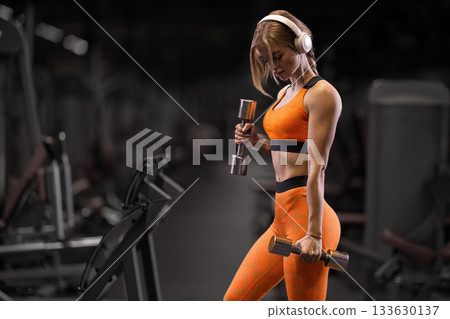 girl in athletic wear performs bicep curls with shiny dumbbells in a fitness gym. She competes her workout while listening to music through her headphones. girl in athletic wear performs bicep curls with shiny dumbbells in a fitness gym. She competes her workout while listening to music through her headphones. 133630137