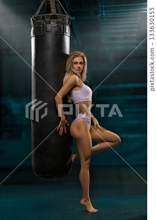 girl with blonde hair stands confidently near a punching bag in a dimly lit gym. She is focused and ready to practice her striking techniques. 133630155