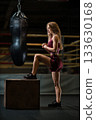 girl stands on a cube in a boxing gym focusing on her balance and strength. The surrounding area features a boxing bag and training equipment. 133630168