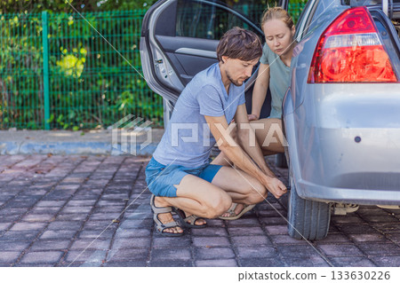 Man loosening a car wheel while woman sits next to him, working together on vehicle maintenance. Concept of teamwork, DIY repair, home car care, safety, and practical skills 133630226