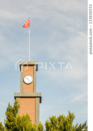 Brick clock tower with flag against blue sky surrounded by trees 133630553