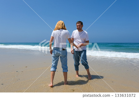 Joyful couple playing at the water's edge on a sunny beach, dressed in casual jeans and white tshirts 133631072