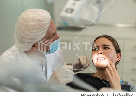 Mature man doctor dentist in white coat examines oral cavity of girl patient in dentistry. 133632999