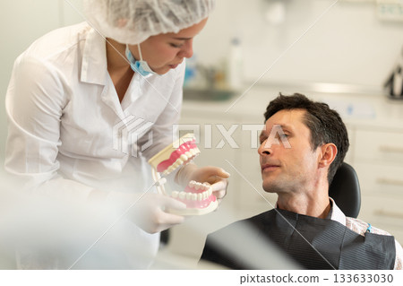 Young woman dentist communicates with male patient, shows model of jaws 133633030