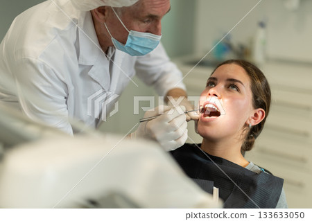 Mature man doctor dentist in white coat examines oral cavity of girl patient in dentistry. 133633050