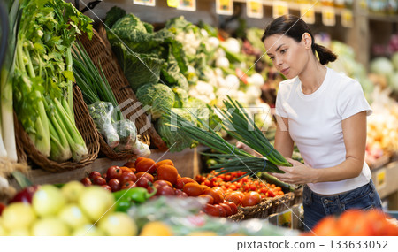 Girl client near vegetable stand in store choose bunch of green onion Girl client near vegetable stand in store choose bunch of green onion 133633052